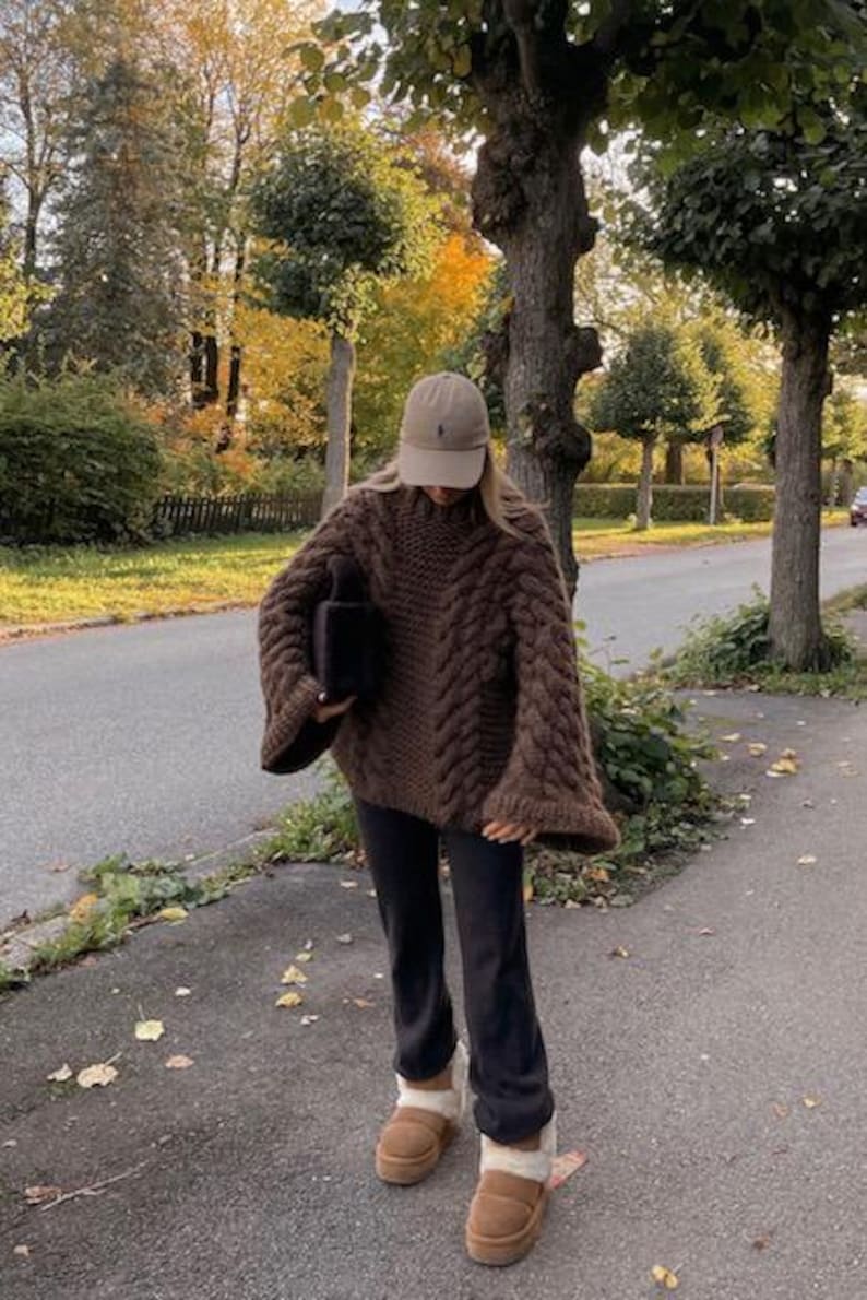 May include: A person wearing a brown cable-knit sweater with wide sleeves, black pants, a tan baseball cap, and brown shearling-lined slippers. The outfit is worn outdoors on a paved path with trees in the background.