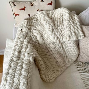 May include: Cream-colored cable knit sweater with a textured design. The sweater is draped over a chair with a dachshund pillow in the background. The sweater has a thick, chunky knit and a cozy appearance.