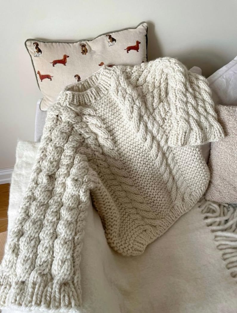 May include: Cream-colored cable knit sweater with a textured design. The sweater is draped over a chair with a dachshund pillow in the background. The sweater has a thick, chunky knit and a cozy appearance.