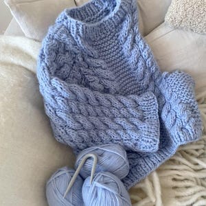 May include: A light blue cable-knit sweater with matching yarn balls and knitting needles. The sweater features a textured cable design and is displayed on a white surface. The yarn balls are light blue and the knitting needles are silver.