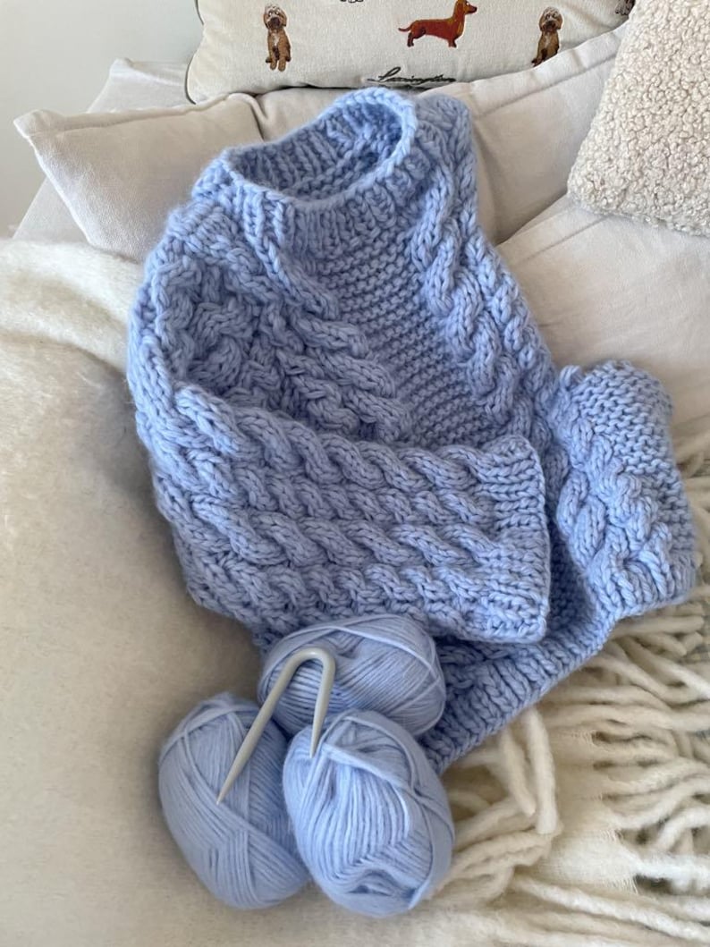 May include: A light blue cable-knit sweater with matching yarn balls and knitting needles. The sweater features a textured cable design and is displayed on a white surface. The yarn balls are light blue and the knitting needles are silver.