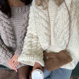 May include: Two cable-knit sweaters, one light gray and one cream, are shown. The sweaters have a chunky knit design. A small brown purse with a strap is visible. One person is holding a white coffee cup.
