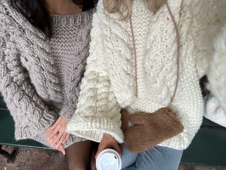 May include: Two cable-knit sweaters, one light gray and one cream, are shown. The sweaters have a chunky knit design. A small brown purse with a strap is visible. One person is holding a white coffee cup.