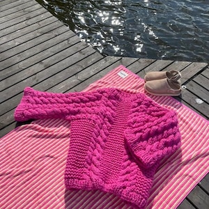 May include: A vibrant fuchsia cable-knit sweater rests on a pink and white striped towel near a body of water. The sweater features a thick, textured design with a crew neck and long sleeves. A pair of light brown slip-on shoes are also visible.