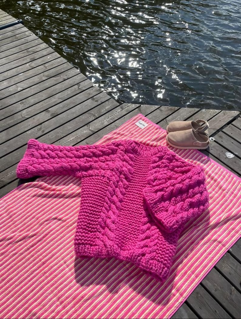 May include: A vibrant fuchsia cable-knit sweater rests on a pink and white striped towel near a body of water. The sweater features a thick, textured design with a crew neck and long sleeves. A pair of light brown slip-on shoes are also visible.