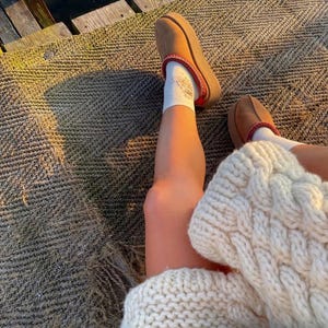 May include: A person wearing brown platform slippers with a red trim and white socks. The person is also wearing a cream-colored cable knit sweater. The scene is set outdoors on a textured surface.