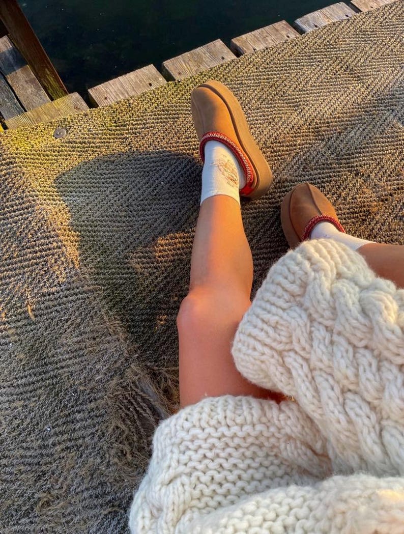 May include: A person wearing brown platform slippers with a red trim and white socks. The person is also wearing a cream-colored cable knit sweater. The scene is set outdoors on a textured surface.