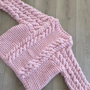 May include: A light pink, hand-knitted sweater with a cable knit design. The sweater features a crew neck and long sleeves with a textured pattern. The garment is made of a soft, chunky yarn, perfect for cooler weather.
