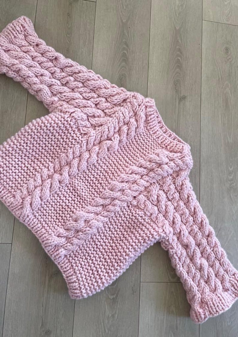 May include: A light pink, hand-knitted sweater with a cable knit design. The sweater features a crew neck and long sleeves with a textured pattern. The garment is made of a soft, chunky yarn, perfect for cooler weather.