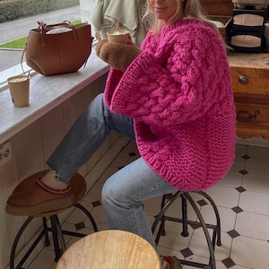 May include: A bright pink, chunky knit sweater with a cable knit pattern. The sweater has long sleeves and is paired with blue jeans. A brown leather tote bag and a cup of coffee are visible on a windowsill.