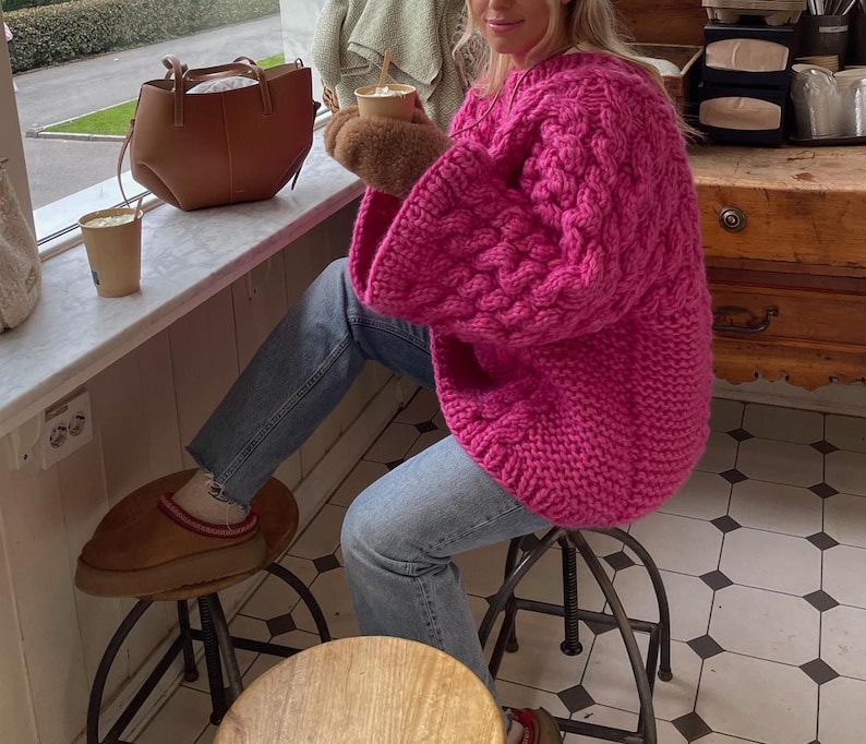 May include: A bright pink, chunky knit sweater with a cable knit pattern. The sweater has long sleeves and is paired with blue jeans. A brown leather tote bag and a cup of coffee are visible on a windowsill.