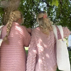 May include: Two people wearing matching light pink knit sweaters with a chunky cable knit design on the sleeves. They are standing under a tree, with one person's hair in a braid and the other with a pink hair clip. They are carrying white tote bags.
