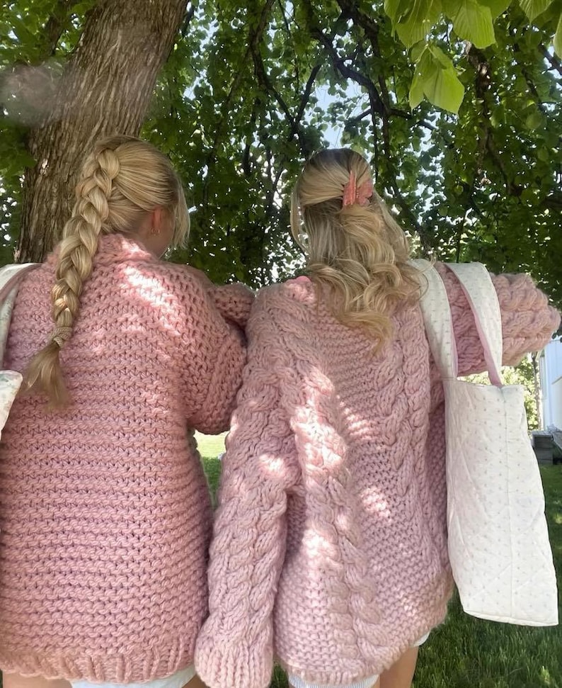 May include: Two people wearing matching light pink knit sweaters with a chunky cable knit design on the sleeves. They are standing under a tree, with one person's hair in a braid and the other with a pink hair clip. They are carrying white tote bags.