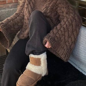 May include: A brown, chunky-knit sweater with wide sleeves and a high neckline. The person is wearing dark gray sweatpants and brown UGG boots with a cream-colored lining. The boots have a brown patch with the brand name.