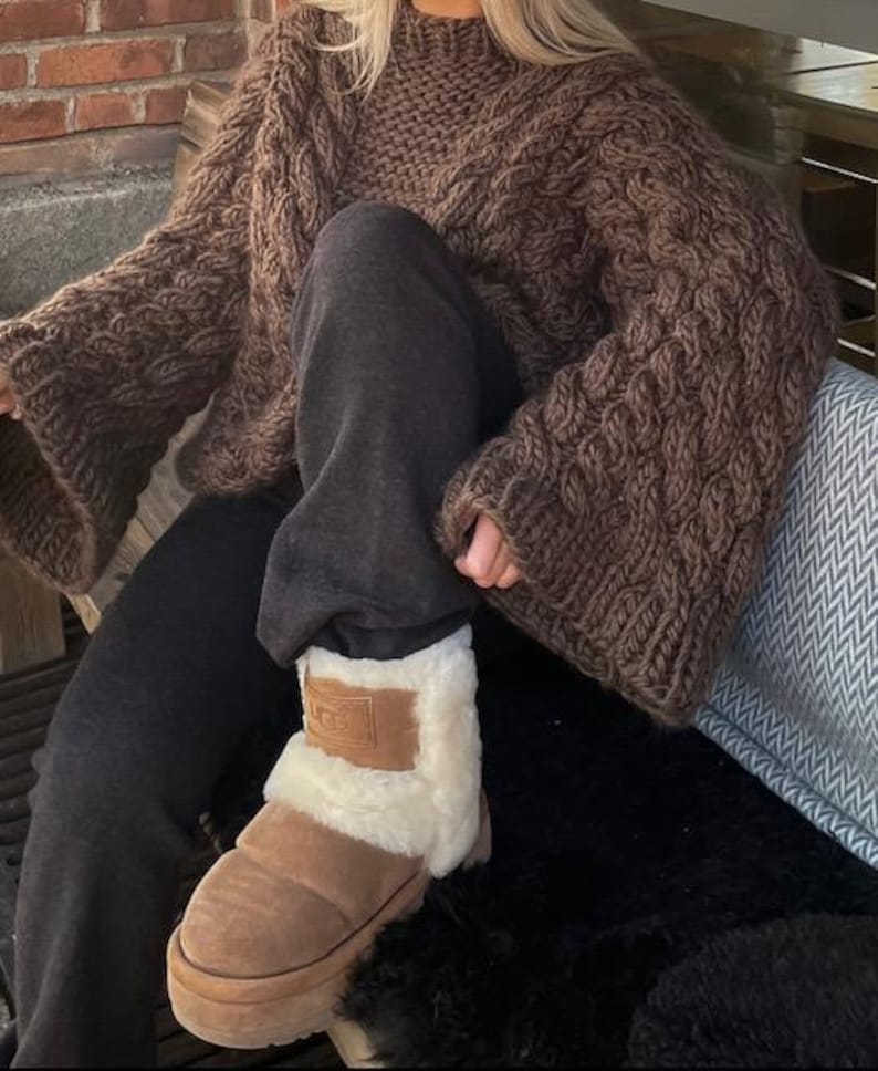 May include: A brown, chunky-knit sweater with wide sleeves and a high neckline. The person is wearing dark gray sweatpants and brown UGG boots with a cream-colored lining. The boots have a brown patch with the brand name.