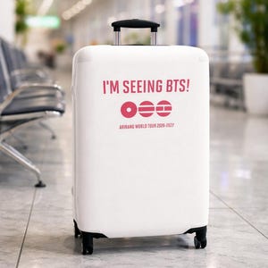 Op de afbeelding: Witte koffer op wielen met de roze tekst "I'M SEEING BTS!". Daaronder een roze cirkel en twee horizontale lijnen. De tekst "ARIRANG WORLD TOUR 2020-2027" staat onderaan. De koffer heeft een zwarte handgreep en wielen.