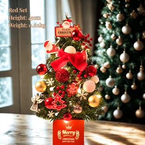 Puede incluir: Un pequeño árbol de Navidad, de unos 45 cm de altura, adornado con adornos rojos, un gran lazo rojo y adornos festivos. La base es una caja roja con el texto "Merry Christmas". El árbol está decorado con luces y adornos festivos.
