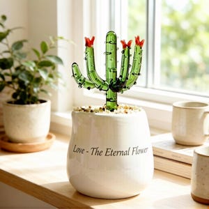 Könnte beinhalten: Ein dekorativer Glas-Kaktus mit roten Blüten in einem weißen Keramiktopf mit Steinen. Der Topf trägt den Text "Love - The Eternal Flower". Der Kaktus steht auf einer Holzoberfläche in der Nähe eines Fensters.