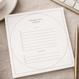 May include: A square notepad with the words "PRIORITIES TODAY" and "TO-DO" printed in black. The notepad has a circular design and lines for writing. A gold pen rests beside the notepad. A cup of coffee and a keyboard are in the background.