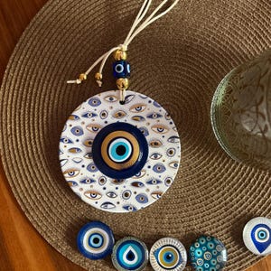 Könnte beinhalten: Runde Wanddekoration mit blau-goldenen Augenmotiven auf weißem Hintergrund. Das Hauptdesign zeigt ein großes zentrales Auge, umgeben von kleineren Augen. Ein Band mit goldenen und blauen Perlen dient zum Aufhängen. Mehrere augenförmige Magnete sind ebenfalls zu sehen.
