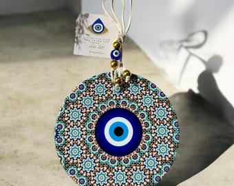 Op de afbeelding: Een ronde, blauwe en witte boze oog-hanger met een gouden kralenketting en een label met de tekst "Bescherming tegen slechte dingen en kwaad lot, Ahlu Art". De hanger is omgeven door een gedetailleerd, ingewikkeld, geometrisch patroon in tinten blauw, groen en bruin.