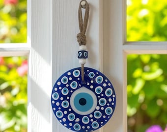 Op de afbeelding: Een blauw en wit Evil Eye-bedel die aan een natuurlijke vezelkoord hangt. De bedel heeft een groot centraal oogontwerp met een blauwe iris en witte accenten. De bedel hangt aan een geknoopt koord met witte en blauwe kralen.