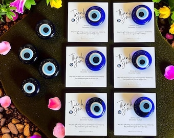 May include: Arrangement of blue and white Evil Eye charms and cards with the words "Thank You" and a date. The charms are round with a blue background, a white iris, and a black pupil. The cards have text about joy and happiness.