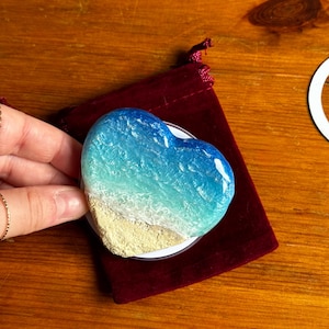 May include: Heart-shaped phone grip depicting a beach scene with blue and turquoise waves, sandy shore, and white details. The grip is on a white base and rests on a burgundy velvet pouch.