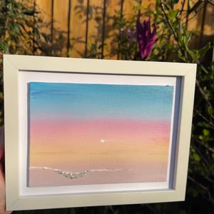 May include: A framed painting of a sunset over a beach with a white frame. The painting is done in shades of pink, orange, yellow, and blue. There are small white dots on the beach.