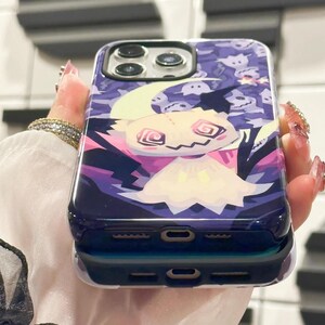 以下が含まれることがあります： 紫と白の携帯電話ケースで、漫画の幽霊のデザインが施されています。ケースは透明で光沢のある仕上げで、カメラの突起が盛り上がっています。