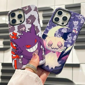 以下が含まれることがあります： ポケモンのキャラクターが描かれた携帯電話ケース2つ。最初のケースは白地に紫と赤のゲンガーのキャラクターと日本語の文字。2つ目のケースは紫地に白と黄色のゴーストのキャラクター
