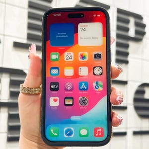 May include: A black iPhone with a colorful home screen. The screen displays the time, date, and various app icons. The phone is being held in a hand with several rings on the fingers.