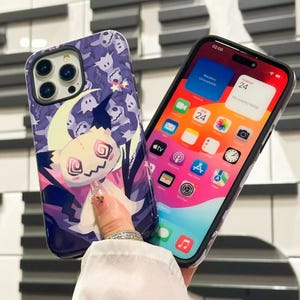 以下が含まれることがあります： 2つのiPhoneケース。1つは、縫い付けられた顔と三日月を持つ漫画のキャラクターを特徴とする紫と黒のデザイン。もう1つは、透明な背面と黒いフレーム。漫画のキャラクターが付いたiPhoneケースには、小さな幽霊のパターンがあります。透明な背面のiPhoneケースには、さまざまなアプリのアイコンが画面に表示されています。