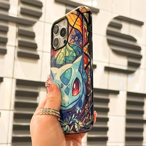 May include: A black phone case with a colorful stained glass design featuring a Pokemon character. The design includes a blue and green creature with red eyes and a yellow background.