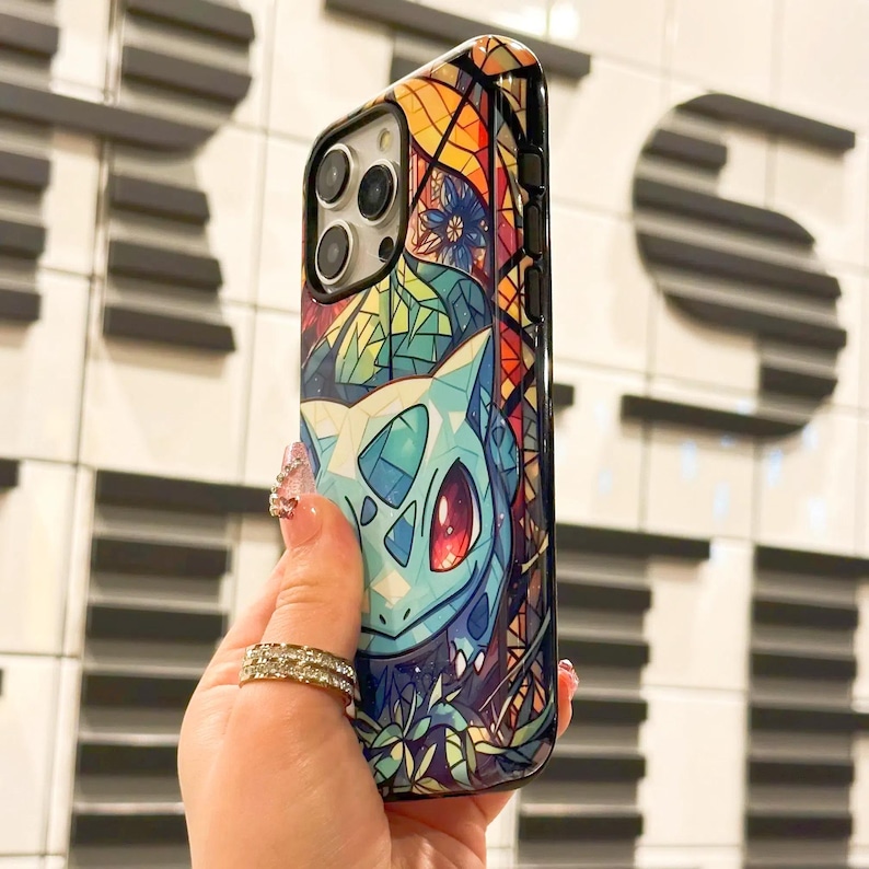 May include: A black phone case with a colorful stained glass design featuring a Pokemon character. The design includes a blue and green creature with red eyes and a yellow background.
