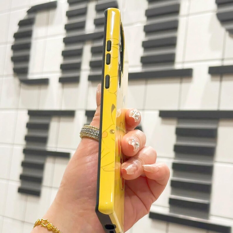 May include: A yellow phone case with a black border and a Pikachu design. The case has a raised edge around the camera lens.