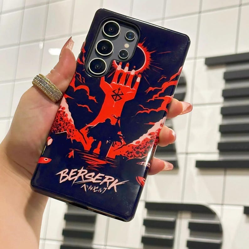 May include: A black phone case with a red and black illustration of a hand reaching up towards a red sun. The case has the text "BERSERK" and "ベルセルク" in white.
