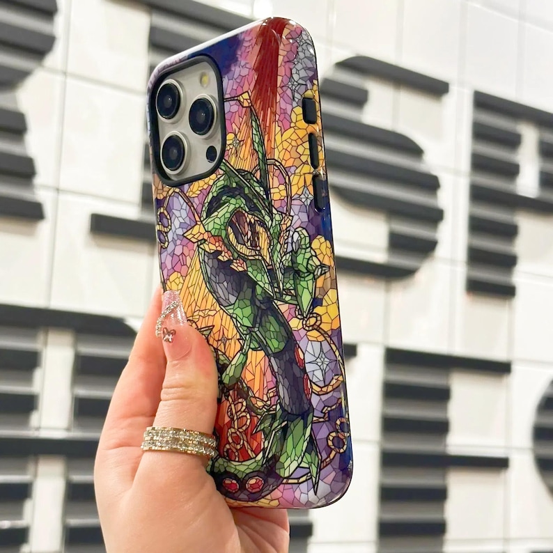 May include: A colorful phone case with a stained glass design featuring a green creature with red eyes. The case is made for an iPhone with a camera lens cutout.