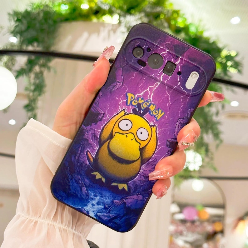 May include: Purple and blue phone case featuring a yellow Psyduck character from Pok&eacute;mon. The case has a lightning bolt design and the word "Pok&eacute;mon" in green. The phone case is held in a hand.