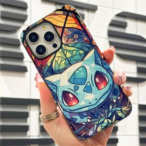 May include: A phone case with a stained glass design featuring a blue and red Pokemon character. The case has a colorful background with a floral pattern.