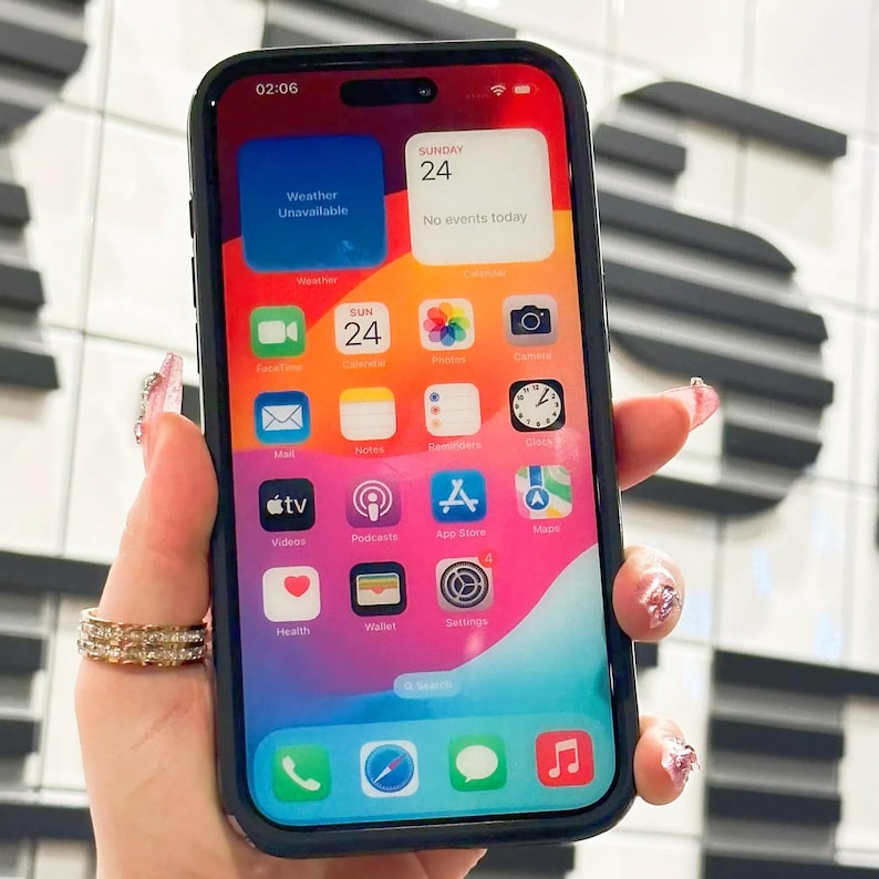 May include: A black iPhone with a bright, colorful home screen. The screen displays the time, date, and various app icons, including Weather, Calendar, Mail, Notes, Reminders, Clock, FaceTime, Camera, Photos, and more. The phone is being held in a hand with several rings on the fingers.