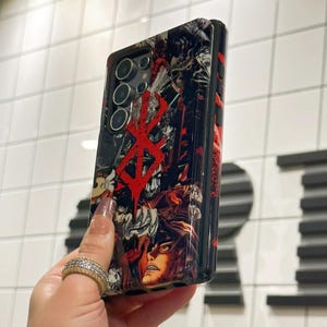 May include: A black phone case with a red and black graphic print of a character from the anime "Berserk". The case has a raised camera bump and a black border.