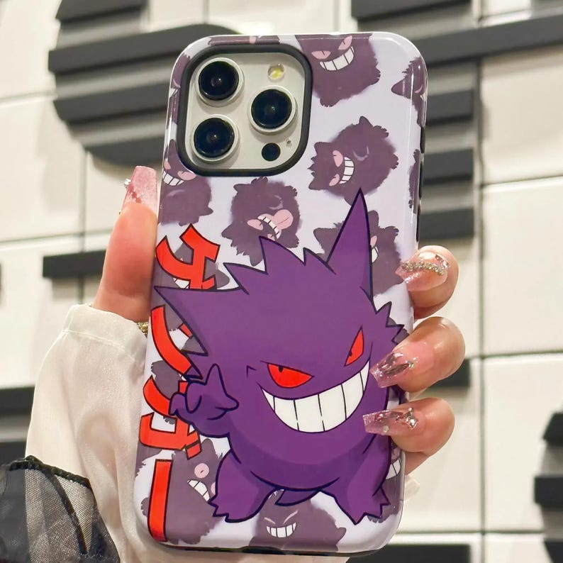 以下が含まれることがあります： 紫と赤のゲンガーポケモンのキャラクターが描かれた白い携帯電話ケース。ケースには、小さなゲンガーのキャラクターのパターンが描かれています。ケースの側面には、赤い文字で日本語が書かれています。