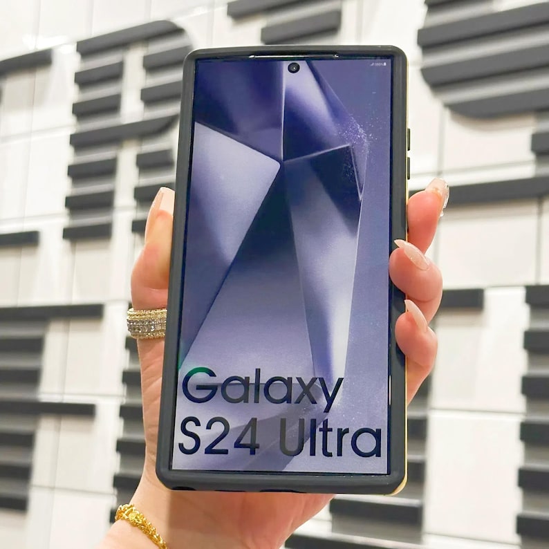 May include: A black smartphone with a gold trim, displaying a purple and gray abstract background on the screen. The phone has the text "Galaxy S24 Ultra" displayed on the screen.