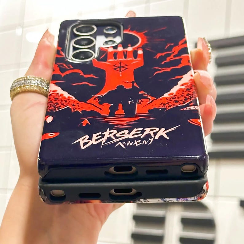 May include: A black phone case with a red and black illustration of a hand with a pentagram and a figure standing in front of it. The text "BERSERK" is written in white with a red outline below the illustration. The case is made for a phone with a triple camera lens.