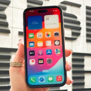 May include: A pink iPhone with a white screen displaying the time 02:06, the date Sunday 24, and a variety of app icons. The phone is being held in a hand with a ring on the finger.