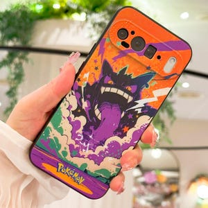 May include: A phone case featuring a colorful design with a cartoon monster character, purple tongue, and the word "Pokemon". The case has an orange top and a black border. The design includes stars and a purple and orange background.