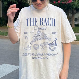 May include: Cream-colored t-shirt with the text "THE BACH Diaries" in navy blue, along with illustrations of a tiara, champagne, and other celebratory items. The text "Me? The Bride? Shut up!" and "NASHVILLE, TN" are also printed on the shirt.