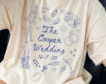 May include: Ivory-colored t-shirt with a blue wedding-themed design. The design includes champagne glasses, rings, and the text "The Cooper Wedding 16-7-25". The shirt has a washed denim design.