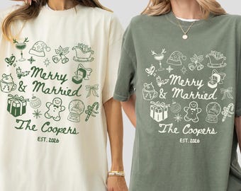 Può includere: Due magliette oversize, una avorio e una verde muschio, ciascuna con una grafica festiva. Il design include il testo "Merry & Married", insieme a illustrazioni a tema festivo, il nome "The Coopers" e l'anno "EST. 2026."